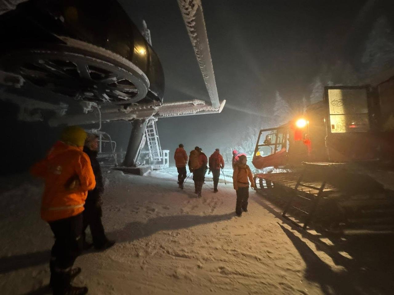 KSAR members in orange jackets walk through falling snow past a frosted chairlift terminal toward a snowcat at night