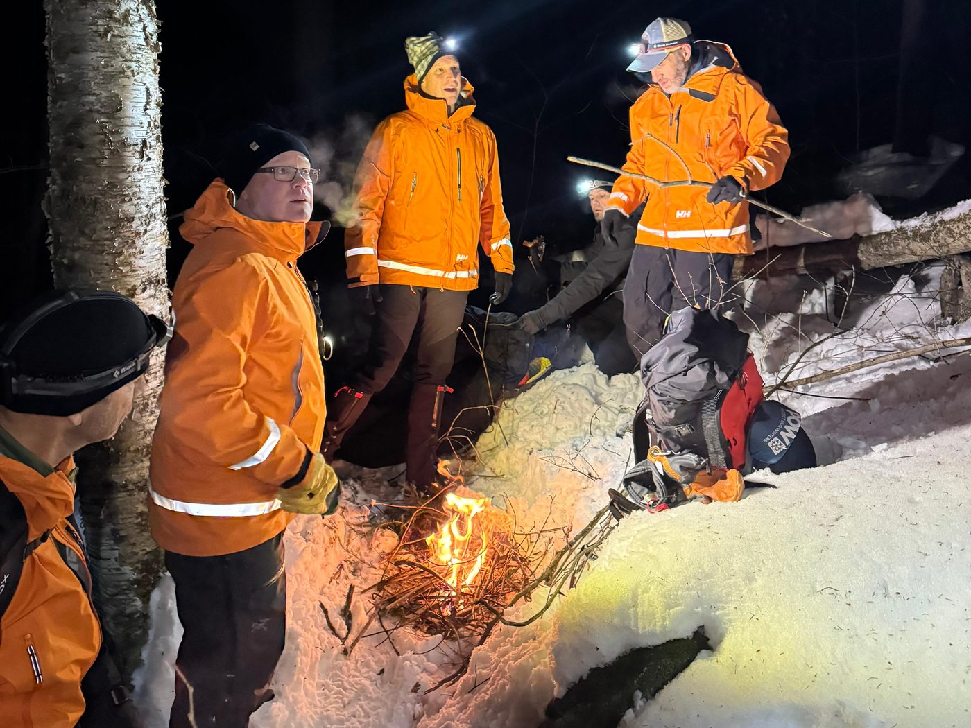 KSAR members in orange jackets and headlamps stand around a small fire in the snowy woods at night