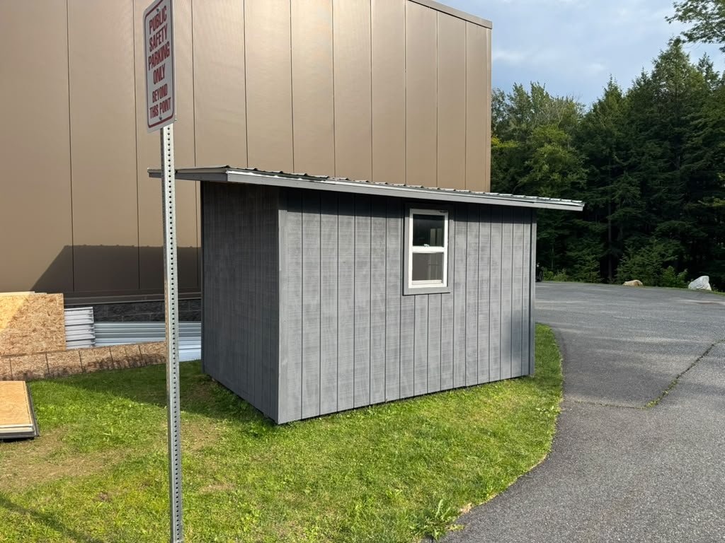 A small gray storage shed with a metal roof and a window, on the grass beside a paved lot
